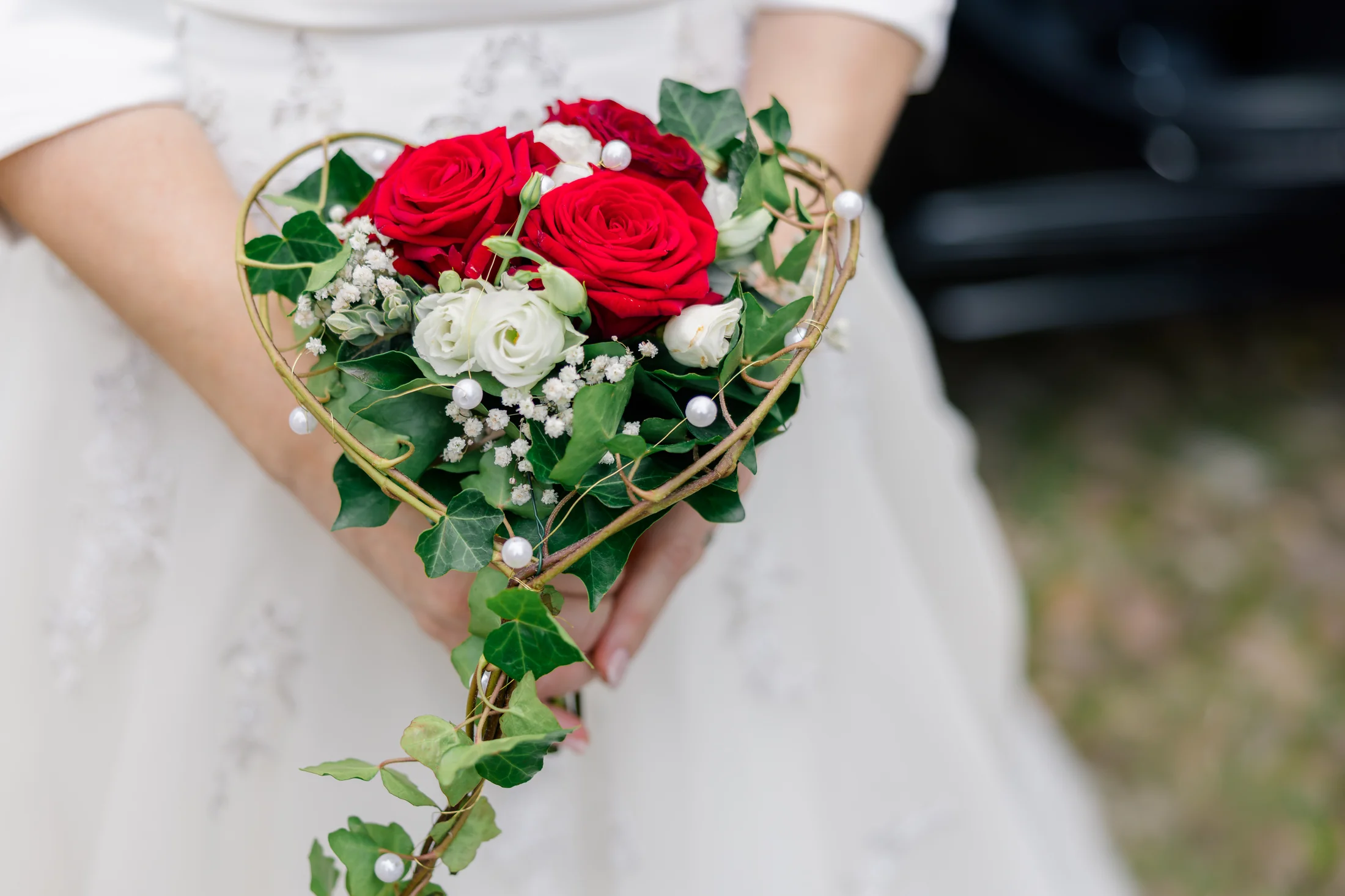 Eine Braut hält einen Strauß aus roten und weißen Rosen mit grünen Blättern vor ihrem Kleid.
